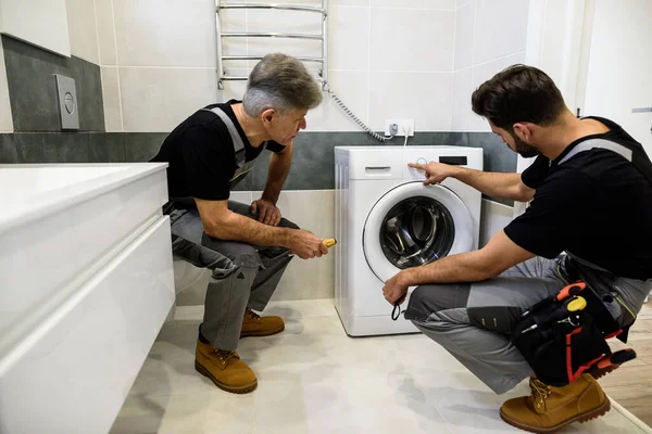 Tiro de longitud completa de dos reparadores, trabajadores en uniforme de trabajo, la lectura de los símbolos de lavado en la mashine lavado antes de fijarlo, la celebración de destornillador en el interior. Servicio de reparación concepto —  Fotos de Stock