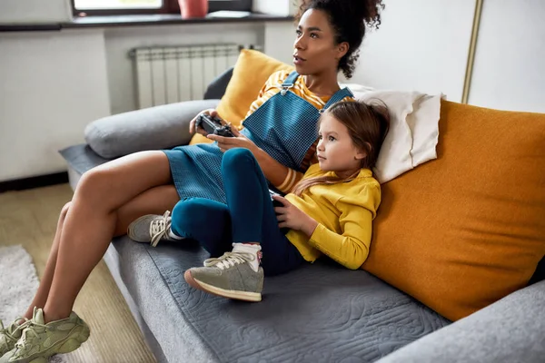 Kul och lek hela dagen. Afrikansk amerikansk kvinna, barnvakt och kaukasisk söt liten flicka som har roligt tillsammans, spelar tv-spel, sitter på soffan. Fritidsaktiviteter, barnpassning — Stockfoto