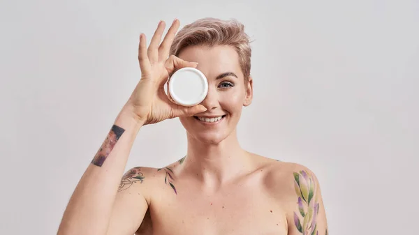 Portrait of beautiful tattooed woman with pierced nose and short hair hiding eye behind white plastic jar of cream or body lotion isolated over grey background — Stock Photo, Image