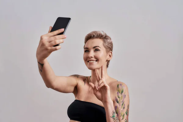 의사소통. 매력적 인 문신을 한 여성이 짧은 머리로 웃으며, 평화의 표시를 하고, 자신의 사진을 찍고, 스마트폰을 사용하여 회색 배경에 고립되어 있다 — 스톡 사진