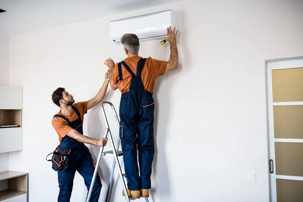 Dos trabajadores en uniforme, maestros de aire acondicionado usando escalera mientras instalan un nuevo aire acondicionado en el apartamento. Concepto de construcción, mantenimiento y reparación —  Fotos de Stock