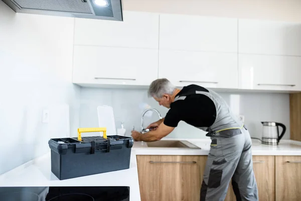 У віці ремонтник в уніформі працює, фіксуючи зламаний кухонний кран з використанням регульованого ключа. Концепція ремонту сервісу — стокове фото