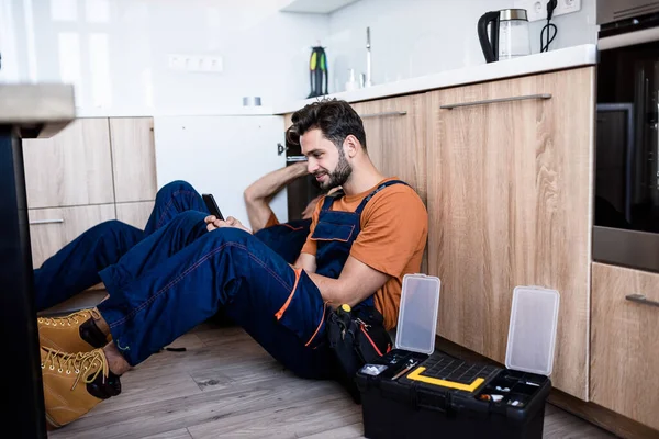 Joven reparador, trabajador en uniforme sentado en el suelo, usando un teléfono inteligente mientras su experimentado colega arregla la tubería del fregadero en el interior. Servicio de reparación, concepto de nueva generación —  Fotos de Stock