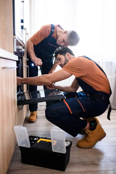 Consigue el mejor servicio de Handyman. Tirador de cuerpo entero de dos reparadores, trabajadores uniformados examinando el horno con destornillador y linterna en el interior. Servicio de reparación concepto —  Fotos de Stock