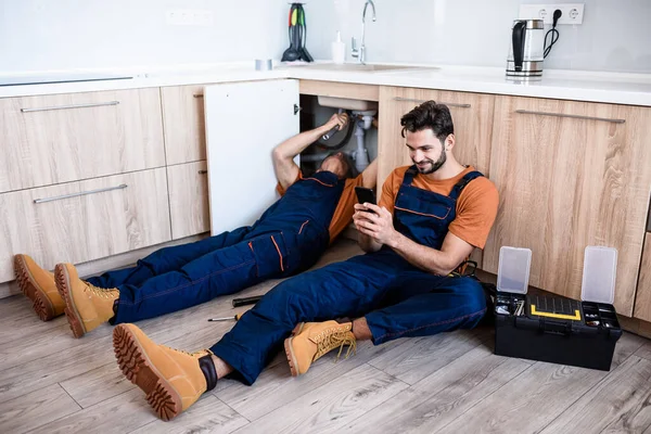 Mladý opravář, dělník v uniformě sedící na podlaze, používající smartphone, zatímco jeho zkušený kolega opravoval umyvadlo. Oprava servisu, koncept nové generace — Stock fotografie