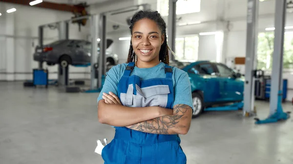 El mejor servicio es mi motivo. Retrato de mujer afroamericana joven, mecánico profesional en uniforme sonriendo a la cámara, de pie en el taller de reparación de automóviles. Servicio de coches, reparación, concepto de mantenimiento — Foto de Stock