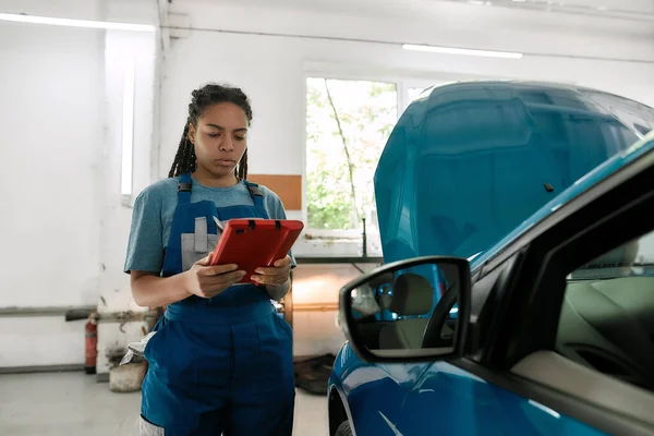 Mujer afroamericana joven, mecánico femenino profesional usando la PC de la tableta, mientras que está parado cerca del coche azul con la capucha abierta en el taller de reparación de automóviles — Foto de Stock