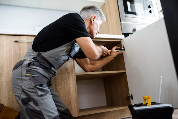 Reparador envejecido en uniforme de trabajo, la fijación de gabinete de cocina utilizando destornillador. Servicio de reparación concepto —  Fotos de Stock