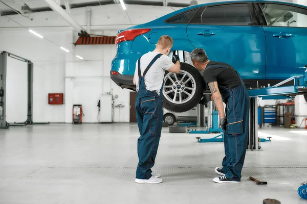 Redefinir el servicio para un coche. tiro de longitud completa de dos mecánicos masculinos que utilizan la antorcha para inspeccionar la rueda del coche levantado en el taller de reparación de automóviles — Foto de Stock