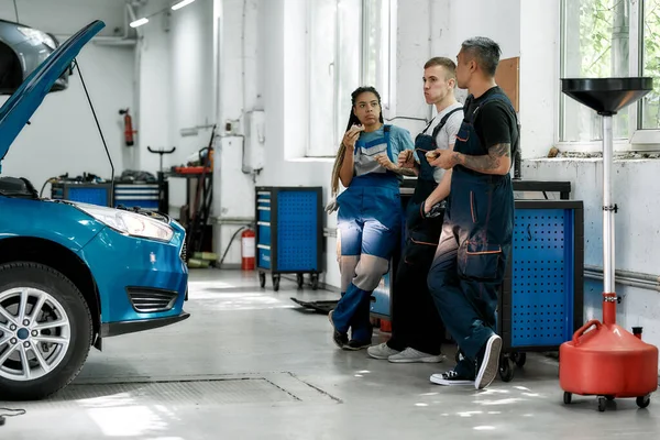 Equipo de diversos mecánicos en uniforme, dos hombres y una mujer hablando y comiendo sándwiches, mientras descansan en el taller de reparación de automóviles — Foto de Stock