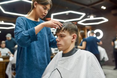 Kuaför servisi. Konsantre olmuş profesyonel berber kız genç yakışıklı adam için modern saç kesimi yapıyor. Berber ya da güzellik salonundaki insanlar.