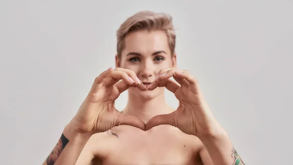 Serce. Close up portret młodej atrakcyjnej pół naga wytatuowana kobieta z idealną skórą patrząc w obiektyw, pokazując serce z rękami odizolowanymi na jasnym tle — Zdjęcie stockowe