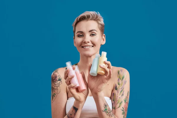 Ritratto di bella donna tatuata con capelli corti che tiene bottiglie di plastica con cosmetici, prodotti per la cura dei capelli e della pelle, isolato su sfondo blu — Foto Stock