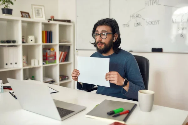 Jeune tuteur en programmation informatique masculin portant des lunettes montrant une feuille de papier vierge à la caméra Web, utilisant un ordinateur portable et donnant des leçons en ligne — Photo