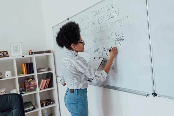 Online iskola. Oldalnézet egy fiatal afro-amerikai tanárnő írja mondat táblára, és tanít angol nyelvet interneten keresztül otthonról — Stock Fotó
