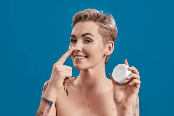 Portrait of beautiful half naked tattooed woman with short hair smiling, while applying cosmetic product on nose, holding jar of cream isolated over blue background — Stock Photo, Image