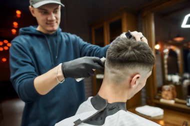 Saç makasıyla çalışan profesyonel berber berber koltuğunda oturan bir genç için modern saç kesimi yapıyor. Bir müşteriye odaklan. Berber dükkanındaki insanlar..