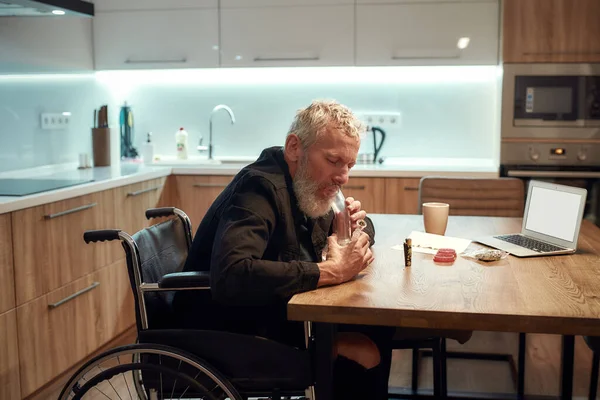 Lätta ditt sinne. Senior handikappade man, konstnär i rullstol håller bong eller glas vattenpipa medan du röker marijuana, skriva sång i köket. Legalisering och beroende av lätta droger — Stockfoto