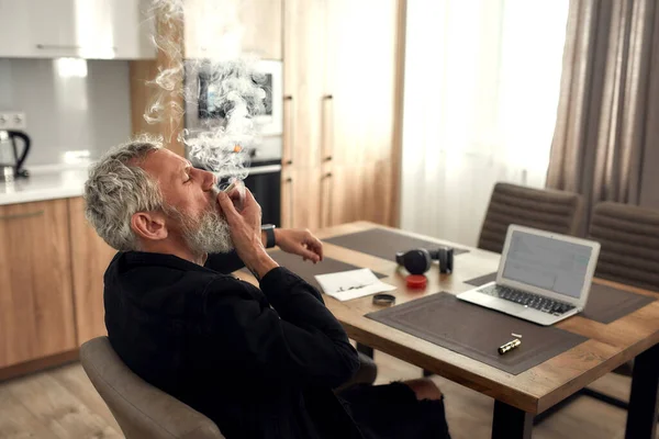 Håll det dåliga humöret borta. Medelålders man, konstnär som röker marijuana cigarett eller joint, sitter i köket och skriver sång med laptop. Marijuanakvarn, tändare, hörlurar och gräs på bordet — Stockfoto
