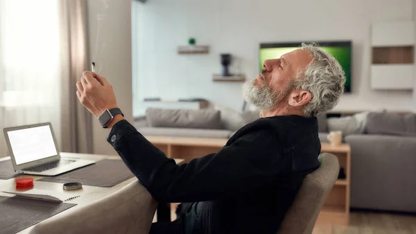 Reste au-dessus. Homme barbu d'âge moyen, artiste fumant une cigarette de marijuana ou un joint, assis à la maison, dans la cuisine et écrivant une chanson à l'aide d'un ordinateur portable. Broyeur de marijuana sur la table — Photo