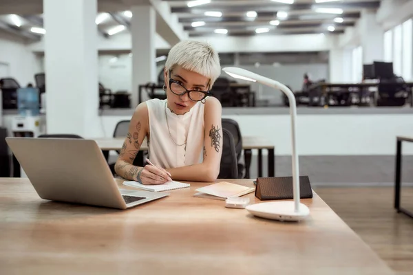 Kemény munkanap. Fiatal, elfoglalt tetovált szőke nő szemüvegben jegyzetel és a kamerába néz, miközben egyedül ül a modern irodában. — Stock Fotó