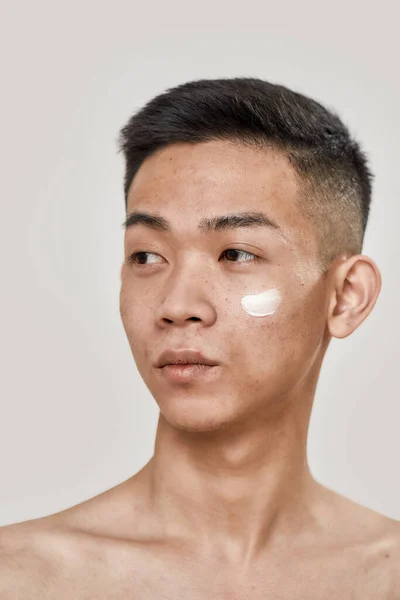 Perfect skincare. Close up portrait of shirtless young asian man with cream applied on his cheek looking aside isolated over white background. Beauty, skincare routine concept — Stock Photo, Image