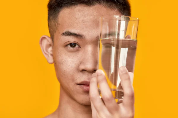 Reequilibrio. Retrato de un joven asiático mirando a la cámara, sosteniendo un vaso de agua cerca de su rostro aislado sobre fondo amarillo. Salud, concepto de bienestar — Foto de Stock