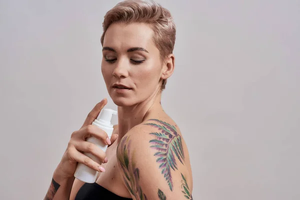Skin with shine. Portrait of beautiful tattooed woman with short hair holding plastic bottle with skin care product, body lotion, applying it on shoulder isolated over grey background — Stock Photo, Image