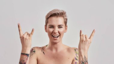 Rock n Roll. Havalı yarı çıplak dövmeli kadın portresi. Kısa saçlı, kameraya gülümseyen, el kol hareketi yapan, arka planda izole edilmiş elleri olan.