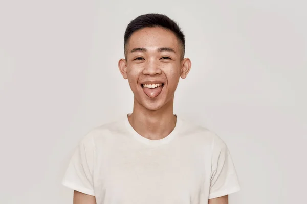 Jen zábava. Portrét veselého mladého Asiata s hladce oholeným obličejem, který vystrkuje jazyk a dívá se na kameru izolovanou na bílém pozadí. Krása, péče o pleť, zdravotní koncepce — Stock fotografie