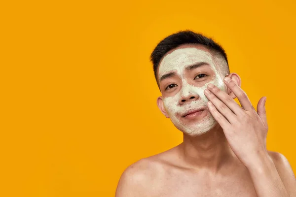 Trate-se. Retrato de alegre sem camisa jovem asiático homem aplicando máscara em seu rosto, olhando para câmera isolada sobre fundo amarelo. Beleza, rotina de cuidados da pele — Fotografia de Stock