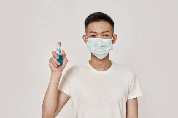 Cuídate. Retrato de un joven asiático con máscara mirando a la cámara, sosteniendo el desinfectante de manos aislado sobre fondo blanco. Concepto de salud —  Fotos de Stock