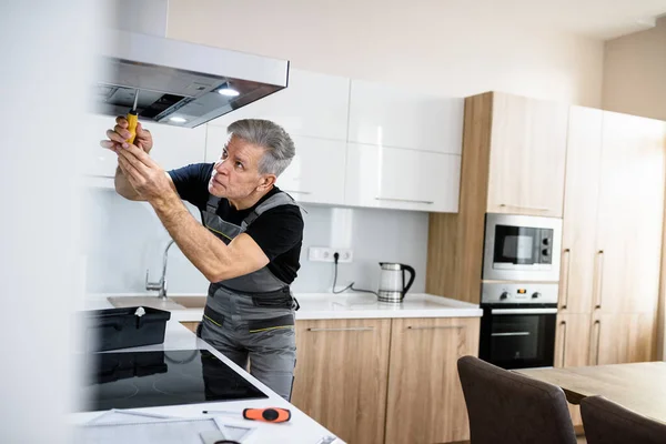 Daha fazla tamir et. Daha dikkatli ol. Üniformalı yaşlı bir tamirci, bozuk mutfak vantilatörünü tornavida kullanarak tamir ediyor. Onarım servisi kavramı — Stok fotoğraf