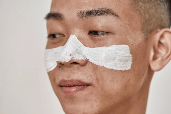 Cuida tu piel. Primer plano retrato de alegre joven asiático hombre con máscara aplicada en su cara mirando a un lado aislado sobre fondo blanco. Belleza, rutina de cuidado de la piel — Foto de Stock