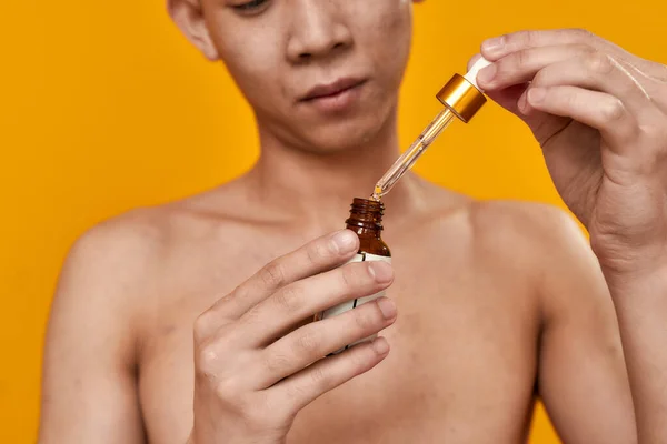 Ragyogjon a bőr. Vágott portré fiatal ázsiai férfi kezében gél szérum üveg pipetta izolált sárga háttér. Szépség, bőr rutin koncepció — Stock Fotó