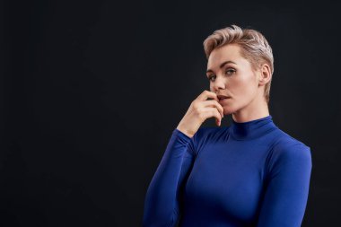 Burnu delinmiş ve boğazlı kısa saçlı çekici bir kadın portresi. Kameraya düşünceli görünüyor. Parmağını koyu arka planda kapalı tutuyor.