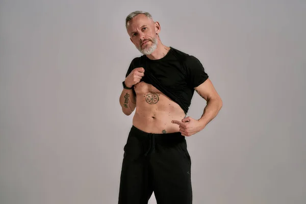 Super Abs. Hombre musculoso de mediana edad levantando camiseta negra mostrando, señalando sus abdominales, tatuajes mientras posa en el estudio sobre fondo gris —  Fotos de Stock