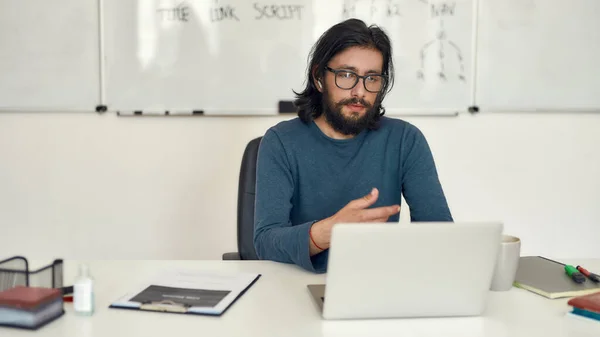 Programming tutor working online. Young focused bearded male teacher teaching computer science online through video call while sitting against whiteboard