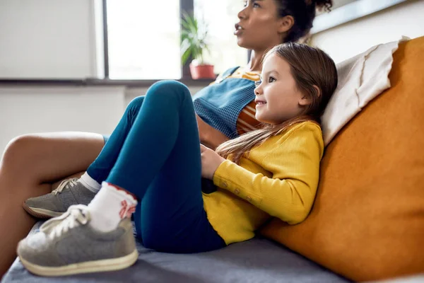 Afrikansk amerikansk kvinna, barnvakt och kaukasisk söt liten flicka som har roligt tillsammans, spelar tv-spel, sitter på soffan — Stockfoto