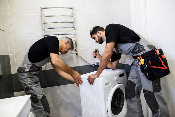 Two repairmen, workers in uniform working, examining washing mashine using flashlight in the bathroom. Repair service concept