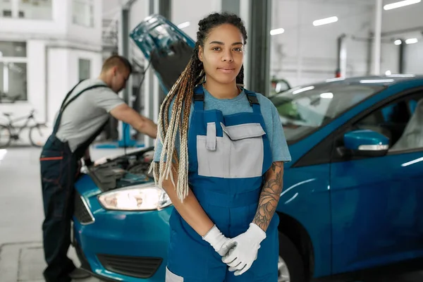 Servicio inteligente. Mujer afroamericana joven, mecánico profesional mirando a la cámara, mientras que su colega masculino reparando el coche en el fondo en el taller de reparación de automóviles — Foto de Stock