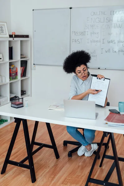 Online tanfolyamot tartok. Fiatal afro-amerikai nő ül a munkahelyén otthon és tanít angol online, elmagyarázza a szabályokat az angol nyelvtan — Stock Fotó