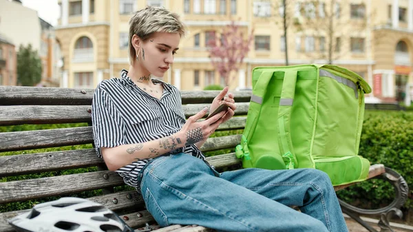 Νεαρή γυναίκα παράδοση με θερμική τσάντα ή σακίδιο ελέγχου προκειμένου χρησιμοποιώντας smartphone, κάθεται στον πάγκο σε εξωτερικούς χώρους. Ταχυμεταφορέας, έννοια της υπηρεσίας παράδοσης — Φωτογραφία Αρχείου
