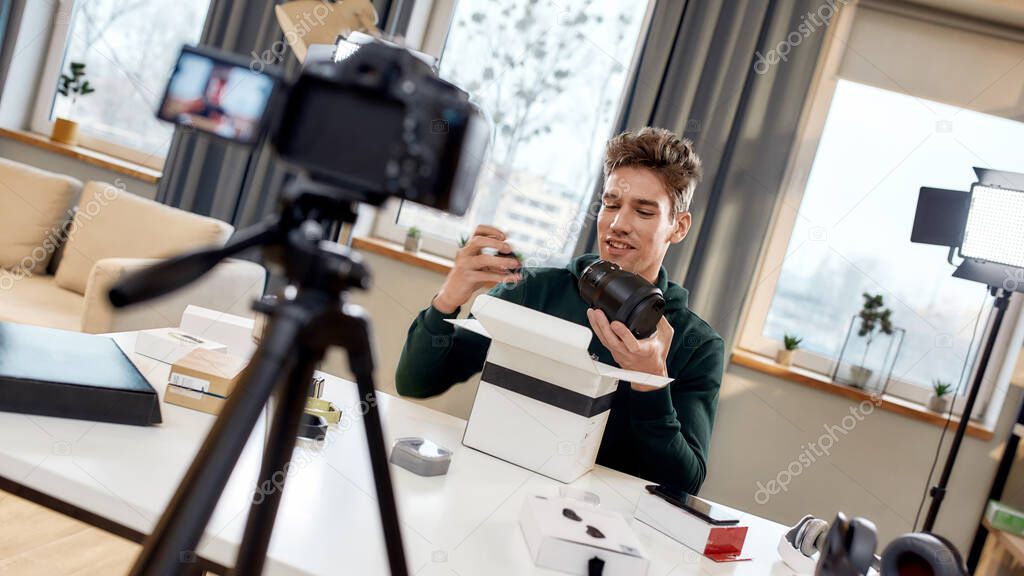 Young male technology blogger recording video blog or vlog about new camera lens and other gadgets at home studio. Blogging, Work from Home concept