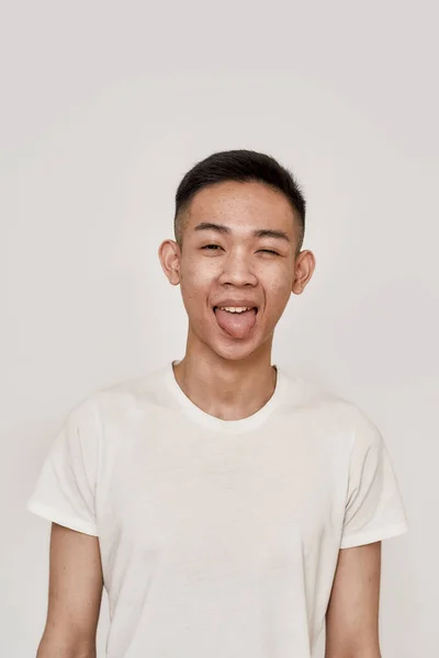 Hravá nálada. Portrét mladého Asiata s hladce oholenou tváří, který vypadá vesele, vystrkuje jazyk a mrká na kameru izolovanou nad bílým pozadím. Krása, péče o pleť, zdravotní koncepce — Stock fotografie