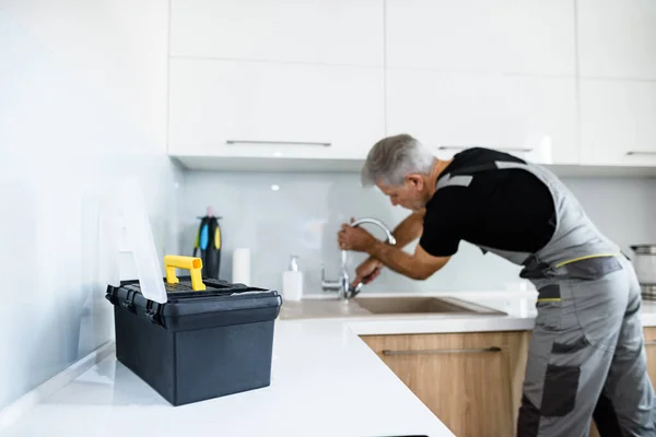 За все. Пожилой ремонтник в униформе работает, чинит сломанный кухонный кран с помощью регулируемого гаечного ключа. Концепция ремонта — стоковое фото
