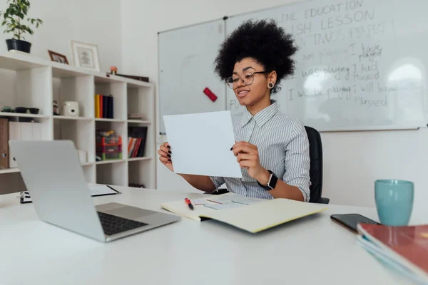 Sorridente afro-americana insegnante di sesso femminile che mostra un foglio bianco di carta alla fotocamera web del suo computer portatile mentre insegna inglese online — Foto Stock