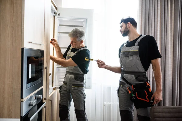 La gente técnica. Reparador de edad en uniforme que fija el refrigerador en la cocina, mientras que su colega le ayuda, trayendo destornillador. Servicio de reparación concepto —  Fotos de Stock