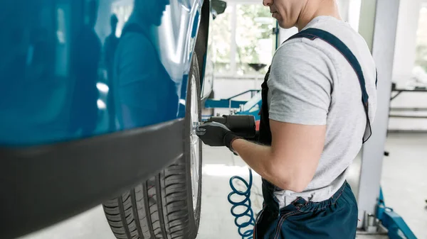 Cropped shot of male mechanic screwing or unscrewing car wheel of lifted automobile by pneumatic wrench at auto repair shop