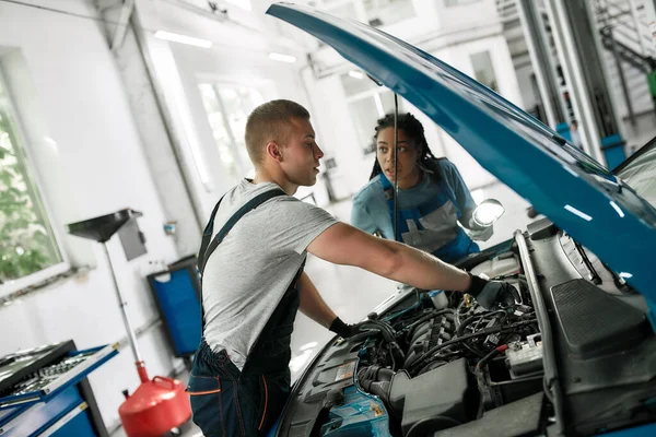 Aprendizaje. Mecánico masculino profesional que repara el motor del coche, aprieta, atornilla con llave mientras que su colega femenino lo mira, sosteniendo la antorcha debajo del capó del coche en la estación de servicio — Foto de Stock